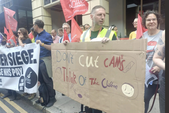 A group of Unite union members and supporters stand on a pavement outside a building holding red Unite flags and protest banners. In the foreground, one person holds a large hand-painted cardboard sign reading, “Don't Cut CAMHS – Think of the Children,” decorated with simple drawings. Another banner calls for an end to the siege of Gaza and a ceasefire. One demonstrator wears a high-visibility vest, while others stand together listening and talking during the protest.