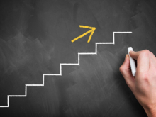 An outline of a staircase drawn in chalk, with an arrow pointing upwards. A hand is holding a stick of chalk.