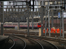 Railway junction with multiple electrified tracks, a London Underground train in the background, and a group of railway workers in orange high-visibility clothing walking beside the tracks.