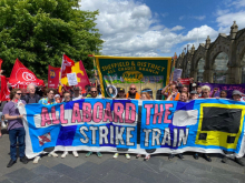 Photo shows a large colourful painted banner reading "All Aboard the Strike Train" leading a demonstration, with an RMT Sheffield branch banner behind it