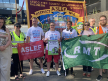 A picket line featuring an Aslef branch banner, with people in the foreground holding a red Aslef flag and a green RMT flag