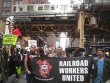 Activists stand underneath a railway bridge holding black Railroad Workers United banner