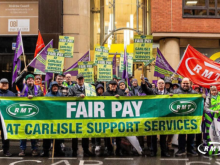 A group of trade union members stand in a city street holding a large green banner reading “FAIR PAY AT CARLISLE SUPPORT SERVICES” with RMT logos on either side. Several people behind them hold matching green and yellow placards with the same message, along with purple union flags and a red RMT flag. The group is gathered outside office buildings on a wet day, wearing coats and hats, appearing to take part in a pay protest or demonstration.