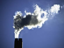 Smoke rises from two industrial chimneys, forming a large dark-and-white plume against a clear blue sky.