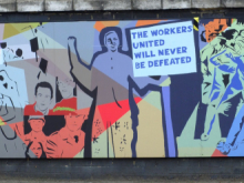 A colorful mural on a wall depicts a group of stylized figures representing workers in protest. One central figure holds a large sign that reads, "THE WORKERS UNITED WILL NEVER BE DEFEATED." The mural features bold colors and abstract shapes, with people of diverse appearances marching together, symbolizing solidarity and labor rights. The central figure is based on Jayaben Desai, the leader of the Grunwick Strike in the 1970s.
