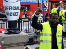 A man wearing a high-visibility yellow vest raises his fist while standing beside a sign reading “RMT Official Picket.” In the background, red double-decker buses and several people are visible on a busy city street.
