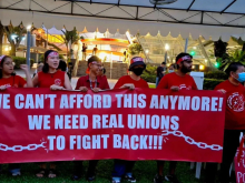 Six individuals holding a red banner with white font. Banner reads “We can’t afford this anymore! We need real unions to fight back!”
