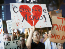 A protester holding a placard reading "Ceasefire Now", superimposed over a broken heart. Other placards are visible in Hebrew.
