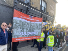 Man standing next to red banner with a crowd of workers holding yellow PCS flags