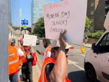 A group of workers wearing bright orange high-visibility vests protest on a city street in daylight. In the foreground, one person holds a handwritten sign reading “Morson & TFL Modern Day Slavery Employer.” Other protesters in the background carry signs calling for better pay and safer working conditions. Tall buildings, parked vehicles, and road signs line the urban street scene under a clear blue sky.