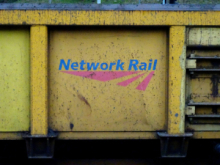 Close-up of a yellow industrial rail vehicle or equipment panel with the blue and red “Network Rail” logo painted on it, showing signs of wear, dirt, and metal fixtures along the edges.