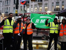 Photo shows an RMT picket line at a London Underground station. Workers make raised-fist salutes in front of a green RMT flag.