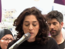 A woman with dark curly hair speaks into a microphone while reading from notes at a public event. Several people stand behind her, including a woman wearing a headscarf and glasses and a bearded man. The background contains blurred banners and display panels.