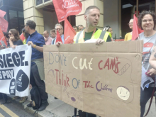 A group of Unite union members and supporters stand on a pavement outside a building holding red Unite flags and protest banners. In the foreground, one person holds a large hand-painted cardboard sign reading, “Don't Cut CAMHS – Think of the Children,” decorated with simple drawings. Another banner calls for an end to the siege of Gaza and a ceasefire. One demonstrator wears a high-visibility vest, while others stand together listening and talking during the protest.