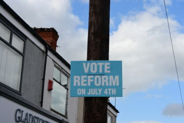A blue campaign poster attached to a wooden pole reads 'VOTE REFORM ON JULY 4TH' in white bold letters. Part of a building with a sign reading 'GLADSTONE' is visible on the left, and the sky with clouds is in the background.