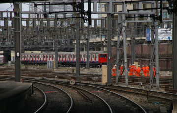 Railway junction with multiple electrified tracks, a London Underground train in the background, and a group of railway workers in orange high-visibility clothing walking beside the tracks.