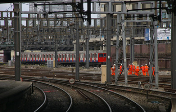 Railway junction with multiple electrified tracks, a London Underground train in the background, and a group of railway workers in orange high-visibility clothing walking beside the tracks.