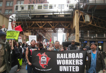 Activists stand underneath a railway bridge holding black Railroad Workers United banner