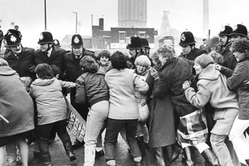 A group of women charging a police line. A colliery is visible on the horizon.