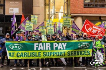 A group of trade union members stand in a city street holding a large green banner reading “FAIR PAY AT CARLISLE SUPPORT SERVICES” with RMT logos on either side. Several people behind them hold matching green and yellow placards with the same message, along with purple union flags and a red RMT flag. The group is gathered outside office buildings on a wet day, wearing coats and hats, appearing to take part in a pay protest or demonstration.