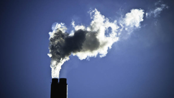 Smoke rises from two industrial chimneys, forming a large dark-and-white plume against a clear blue sky.
