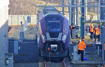 Purple East Midlands Railway train at a test or maintenance facility, with several railway workers in orange high-visibility clothing standing beside the tracks and equipment.