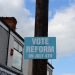 A blue campaign poster attached to a wooden pole reads 'VOTE REFORM ON JULY 4TH' in white bold letters. Part of a building with a sign reading 'GLADSTONE' is visible on the left, and the sky with clouds is in the background.