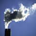 Smoke rises from two industrial chimneys, forming a large dark-and-white plume against a clear blue sky.