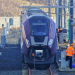 Purple East Midlands Railway train at a test or maintenance facility, with several railway workers in orange high-visibility clothing standing beside the tracks and equipment.