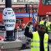 A man wearing a high-visibility yellow vest raises his fist while standing beside a sign reading “RMT Official Picket.” In the background, red double-decker buses and several people are visible on a busy city street.