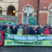 A group of railway workers and union supporters gather outside a station entrance, holding a large green banner reading “No Driver-Only Operation on East West Rail” alongside RMT and regional union banners. The crowd includes people in winter clothing, with a station’s brick and stone façade behind them.