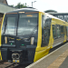 Yellow-and-black Merseyrail train standing at a station platform, displaying "Kirkby" as its destination, with a footbridge spanning the tracks in the background.