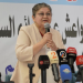 A woman wearing glasses and a light beige blazer (Yanar Mohammed) speaks into a handheld microphone at a press conference. She stands behind a table with several broadcast microphones in front of her, including ones labeled with TV channel logos. Arabic text is visible on the backdrop behind her.