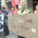 A group of Unite union members and supporters stand on a pavement outside a building holding red Unite flags and protest banners. In the foreground, one person holds a large hand-painted cardboard sign reading, “Don't Cut CAMHS – Think of the Children,” decorated with simple drawings. Another banner calls for an end to the siege of Gaza and a ceasefire. One demonstrator wears a high-visibility vest, while others stand together listening and talking during the protest.